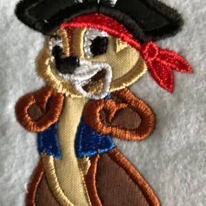 May include: Embroidered patch of a cartoon chipmunk wearing a black pirate hat with a white skull and crossbones design, a red bandana, and a blue waistcoat. The chipmunk is smiling and has brown fur.