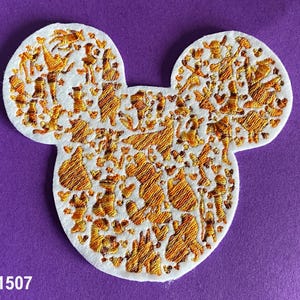 May include: A white felt Mickey Mouse head shape with an embroidered design of various characters from Disney movies in gold thread.