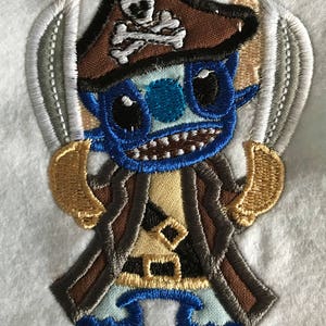 May include: Embroidered patch of a blue, brown and gold cartoon character wearing a pirate hat with a skull and crossbones. The character is holding two swords.