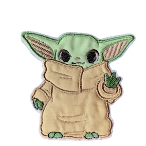 Op de afbeelding: Een geborduurde patch met een populair fictief personage, bekend als "Baby Yoda", uit de Star Wars-franchise. Het personage is afgebeeld in een bruine robe en heeft grote, donkere ogen.