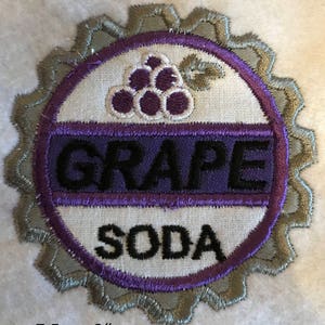 Puede incluir: Un parche bordado con forma de tap&oacute;n de botella con las palabras "GRAPE SODA" y un dise&ntilde;o de uva morada. El parche tiene un di&aacute;metro de aproximadamente 7,5 cm.