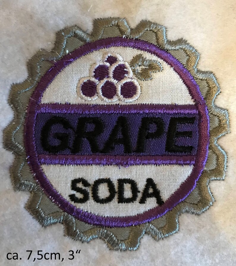 Puede incluir: Un parche bordado con forma de tap&oacute;n de botella con las palabras "GRAPE SODA" y un dise&ntilde;o de uva morada. El parche tiene un di&aacute;metro de aproximadamente 7,5 cm.