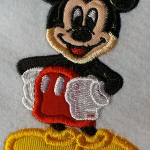 May include: Embroidered patch of Mickey Mouse, featuring a black outline, red shorts, yellow shoes, and a white background.