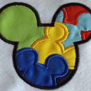 Könnte beinhalten: Ein farbenfroher Filz-Applikations-Puzzle in Form eines Mickey-Mouse-Kopfes. Die Puzzleteile sind grün, gelb, blau und hellblau mit roten Nähten. Die Teile sind zusammengenäht, um die Form des Mickey-Mouse-Kopfes zu bilden.