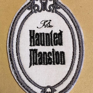 Może przedstawiać: Biała łatka z tkaniny z czarnym haftem. Łatka ma owalny kształt z ozdobną ramką i tekstem "The Haunted Mansion" w stylu gotyckim.