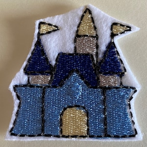 Peut inclure: Écusson de château brodé. Le château bleu clair a des toits bleu foncé et des tours argentées. Le patch est sur un fond en feutre blanc avec des coutures noires. Deux drapeaux dorés sont au sommet du château.