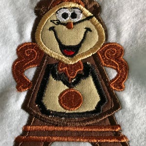 May include: Embroidered patch of Cogsworth, the animated clock from Disney's Beauty and the Beast. He is smiling with a brown and gold clock face and a brown body.