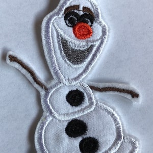 Könnte beinhalten: Weißer Filz-Aufnäher von Olaf aus dem Film Frozen. Olaf ist ein Schneemann mit einer Karottennase, schwarzen Augen und einem braunen Zweig für Arme. Er lächelt mit offenem Mund.