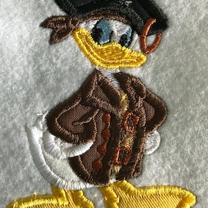 May include: Embroidered patch of a cartoon duck wearing a brown jacket and a black pirate hat with a skull and crossbones. The duck has a yellow beak and white feathers.