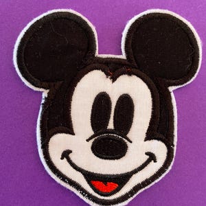 Könnte beinhalten: Ein gestickter schwarzer und weißer Aufnäher mit dem Gesicht von Mickey Mouse, dessen Mund rot ist. Der Aufnäher befindet sich auf einem violetten Hintergrund.
