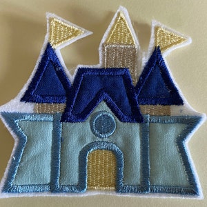 May include: A blue and white felt castle applique with a yellow tower and flags. The castle is outlined in blue and has a white background.