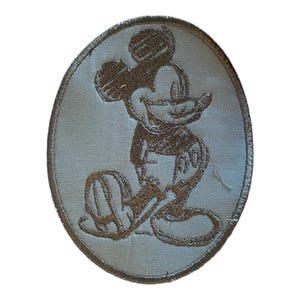 May include: A white oval-shaped patch with a brown embroidered image of Mickey Mouse. Mickey is standing with his right hand on his hip and his left foot forward.