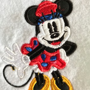 May include: Embroidered patch of a cartoon mouse wearing a red and blue hat and a red dress with white polka dots. The mouse is smiling and has black eyes and a black nose.