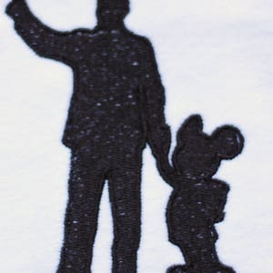 May include: Black embroidered silhouette of a person pointing to the right, with a smaller silhouette of Mickey Mouse standing to the right of the person.