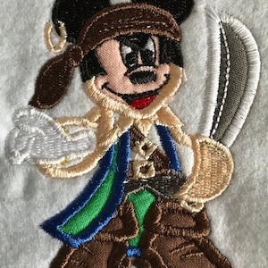 Puede incluir: Parche bordado de Mickey Mouse vestido de pirata. Lleva un sombrero marrón, un chaleco marrón, una faja azul y verde y botas marrones. Sostiene una espada en su mano derecha.