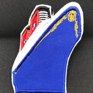 May include: An iron-on patch embroidered with a blue and white ship, featuring a red and white striped smokestack. The ship has a gold trim around the top and a white outline. The patch measures approximately 4 inches by 3 inches.