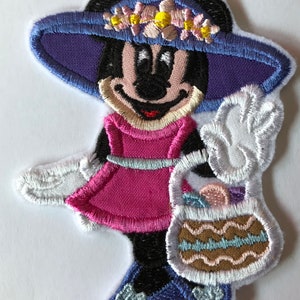 May include: An embroidered patch featuring a cartoon Minnie Mouse wearing a purple hat with flowers, a pink dress, and white gloves. She is holding a brown and blue basket with a white handle.