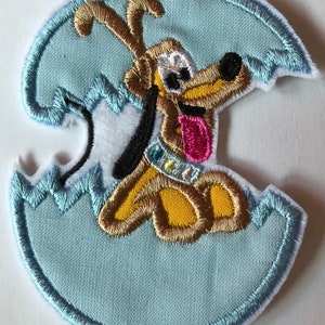 May include: A blue and white embroidered patch featuring a cartoon dog with brown fur, a pink tongue, and a colourful collar, peeking out of a cracked egg.