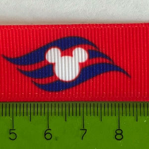 Könnte beinhalten: Rotes Band mit einem sich wiederholenden Muster aus einem weißen Mickey Mouse-Kopf in einem blau-weißen stilisierten Wellendesign. Das Band wird mit einem grünen Lineal in Zentimetern gemessen.