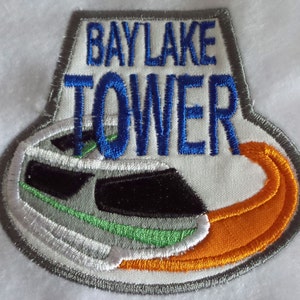 May include: A white embroidered patch with the text "BAYLAKE TOWER" in blue lettering. The patch features a stylised image of a boat with orange, green and grey details.