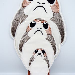 Könnte beinhalten: Gestickte Aufnäher mit drei Porg-Charakteren aus Star Wars, die übereinander gestapelt sind. Die Aufnäher sind weiß mit braunen und schwarzen Details, einschließlich großer Augen und einem finsteren Gesichtsausdruck. Der untere Aufnäher hat braune Füße.