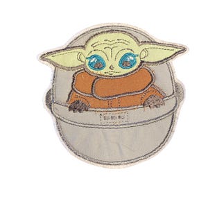 May include: Iron-on patch featuring an embroidered green alien character with large eyes, wearing an orange robe, sitting in a grey pod.