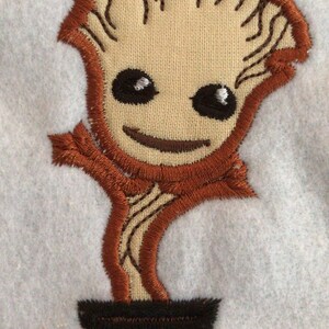 May include: Embroidered patch of a cartoon tree-like character. The character has a tan face, brown hair, and a smiling expression. It is in a brown pot. The patch is on a light blue background.