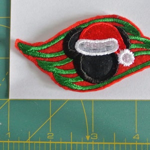 May include: Red and green embroidered patch of a cartoon mouse wearing a Santa hat. The mouse has black ears and a black face. The patch is shaped like a leaf.