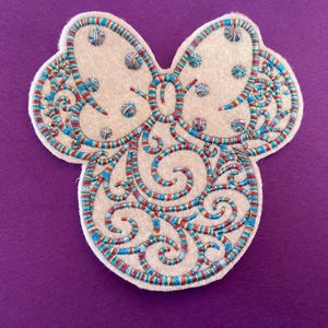 May include: A white felt applique with a blue, red and gold embroidered design. The design is a stylised floral pattern with a bow at the top.