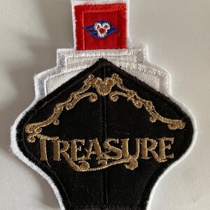 May include: A black and white embroidered patch with a gold "TREASURE" inscription. The patch features a white outline and a red square with a blue Disney cruise ship logo on top.