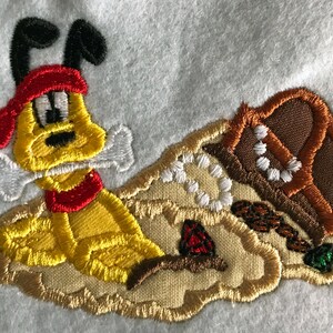 May include: Embroidered patch of a yellow dog wearing a red bandana and holding a white bone. The dog is on a tan background with a brown treasure chest and white pearls.