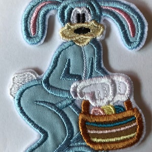 May include: A blue bunny wearing a blue jumpsuit holds a brown basket filled with colourful Easter eggs. The bunny has pink ears and a yellow face.