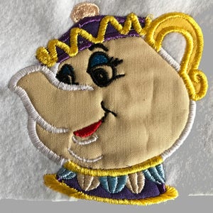May include: Embroidered patch of a cartoon teapot with a yellow, purple, and blue crown, a yellow spout, and a white, yellow, and red smiling face.