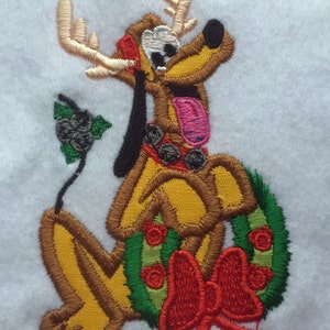 May include: Embroidered Christmas design featuring a brown and yellow dog with antlers wearing a red and green wreath. The dog is sitting with its tongue sticking out and has a pink nose.