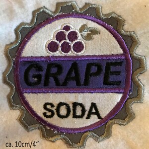 Puede incluir: Un parche bordado con forma de tap&oacute;n de botella con las palabras "GRAPE SODA" y un dise&ntilde;o de uva morada en hilo morado y gris.