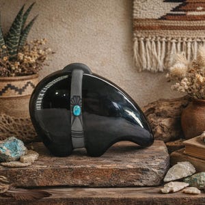 May include: A smooth, black, bear-shaped ceramic sculpture featuring a grey band across its midsection. The band is decorated with a small, oval, blue turquoise stone and subtle etched line patterns, creating a distinct contrast against the glossy black finish of the bear's body.