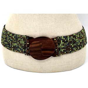 May include: A wide belt featuring a woven band of small, multi-colored beads in shades of lime green, dark brown, and black. The center of the belt is secured by a large, circular, dark brown wooden buckle with a smooth, polished wood grain finish.