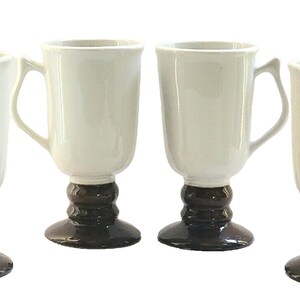 May include: Four white ceramic mugs with dark brown pedestal bases and handles. The mugs have a classic design, suitable for coffee or tea. The mugs are arranged in a row against a white background.