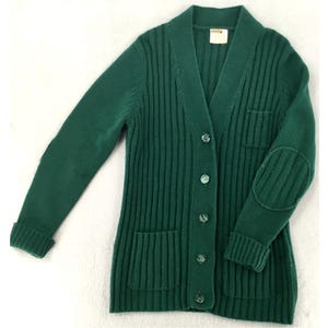 May include: A forest green long-sleeve cardigan featuring a ribbed knit texture, a shawl collar, and a button-front closure. The garment includes two front patch pockets and reinforced oval elbow patches on the sleeves, with a white brand label.