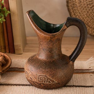 May include: A brown ceramic pitcher with a dark handle and a green interior. The pitcher has a textured surface and a decorative design near the base. The pitcher is on a beige and brown striped cloth.
