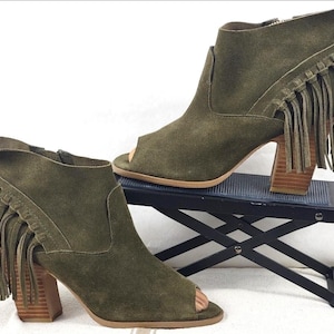 May include: Olive green suede peep-toe ankle boots with fringe detailing. The boots have a block heel and a zipper closure. The shoes are displayed on a small black stand.