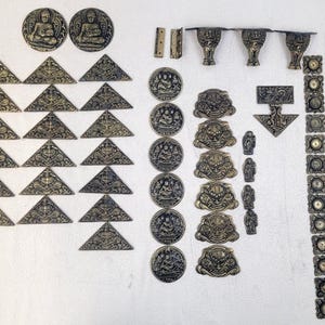 May include: An assortment of antique bronze-colored metal decorative hardware pieces, including door knockers, triangular corner pieces, round medallions with seated figures, and various other ornate embellishments. The collection is arranged on a white surface.