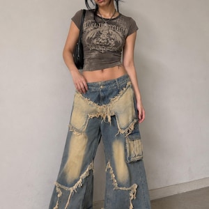 May include: A dark brown short-sleeve crop top with a graphic print and text, paired with wide-leg blue denim jeans featuring large beige distressed patches, frayed edges, and a side cargo pocket, worn with black platform shoes.