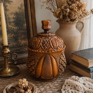 May include: A round, amber-colored glass jar with a matching lid, featuring a detailed pressed pattern of vertical panels, diamond cross-hatching, and starburst motifs. The glass container has a thick, textured surface and a knob-style handle.