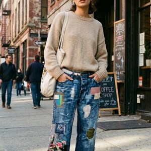 May include: A beige crewneck knit sweater paired with blue denim jeans featuring various fabric patches, distressed details, and frayed hems. The outfit includes a white canvas tote bag worn over the shoulder and brown leather ankle boots.