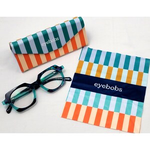 May include: Eyebobs brand eyeglasses with a dark blue and teal tortoiseshell frame, accompanied by a matching triangular hard case and a square microfiber cleaning cloth. Both the case and the cloth feature a vibrant striped pattern in orange, teal, and light blue, with the white eyebobs logo printed on each item.