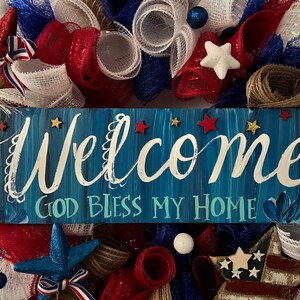 K&ouml;nnte beinhalten: Eine patriotische Girlande in Rot, Wei&szlig; und Blau. Sie zeigt ein blaues Holzschild mit dem Wort "Welcome" in wei&szlig;er Schrift und dem Satz "God Bless My Home". Verziert mit Sternen und B&auml;ndern, die ein festliches Aussehen erzeugen.