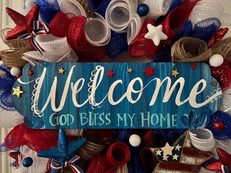 K&ouml;nnte beinhalten: Eine patriotische Girlande in Rot, Wei&szlig; und Blau. Sie zeigt ein blaues Holzschild mit dem Wort "Welcome" in wei&szlig;er Schrift und dem Satz "God Bless My Home". Verziert mit Sternen und B&auml;ndern, die ein festliches Aussehen erzeugen.