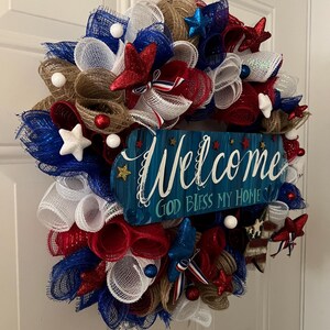 K&ouml;nnte beinhalten: Ein patriotischer Kranz mit roten, wei&szlig;en und blauen Netzb&auml;ndern, Sternen und dekorativen Akzenten. Ein blaues Holzschild in der Mitte tr&auml;gt die Aufschrift "Welcome, God Bless My Home."