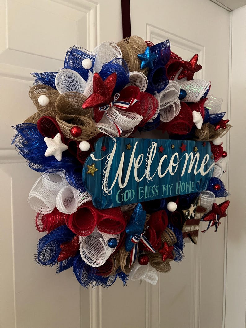 K&ouml;nnte beinhalten: Ein patriotischer Kranz mit roten, wei&szlig;en und blauen Netzb&auml;ndern, Sternen und dekorativen Akzenten. Ein blaues Holzschild in der Mitte tr&auml;gt die Aufschrift "Welcome, God Bless My Home."